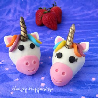 two white chocolate strawberry unicorns set on a purple and pink watercolor background with fresh strawberries set behind