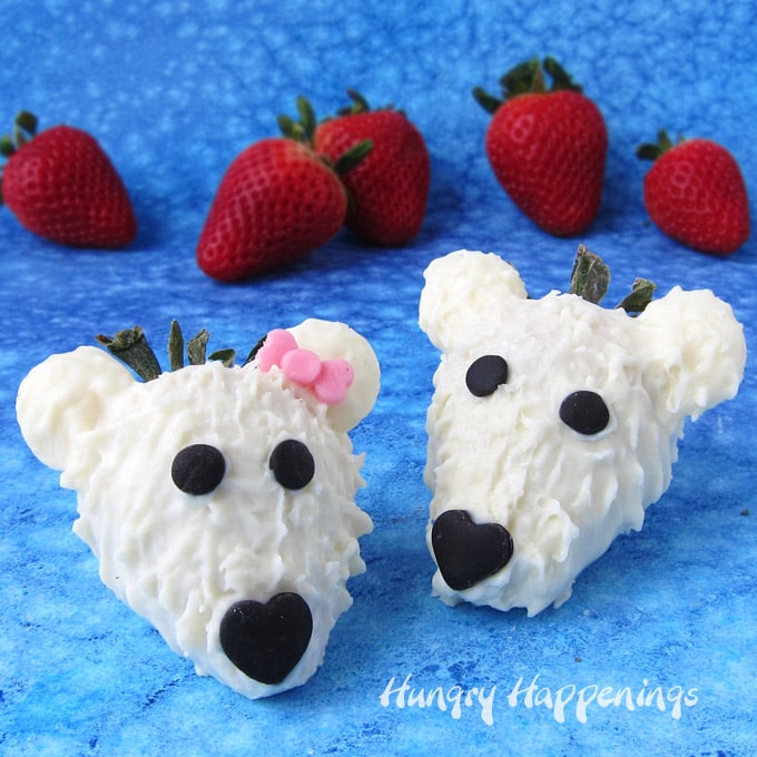two white chocolate dipped strawberries are decorated with modeling chocolate to look like polar bears and are set on a blue watercolor background with strawberries in the background