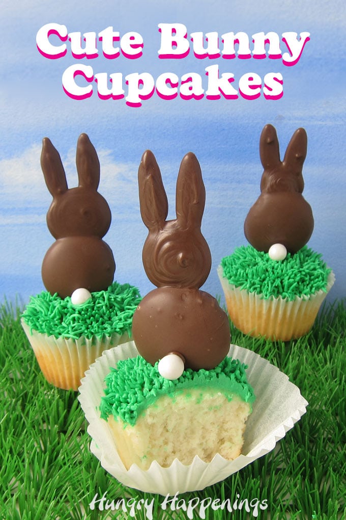three cute bunny cupcakes are displayed on plastic grass set in front of a blue sky backdrop. Each white cupcake is topped with green frosting grass and one chocolate bunny. 