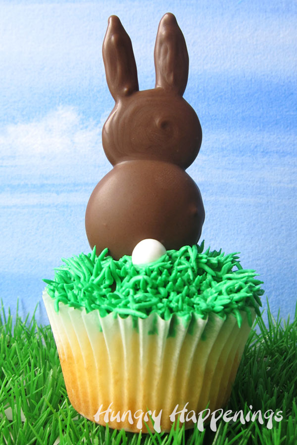 A chocolate bunny with a white candy tail is siting on top of a frosting grass covered cupcake on a grassy patch set in front of a blue sky background