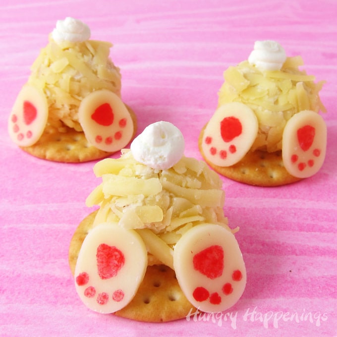 3 mini cheese ball bunnies with cream cheese tails and white cheddar cheese feet are set on round crackers displayed on a pink watercolor background