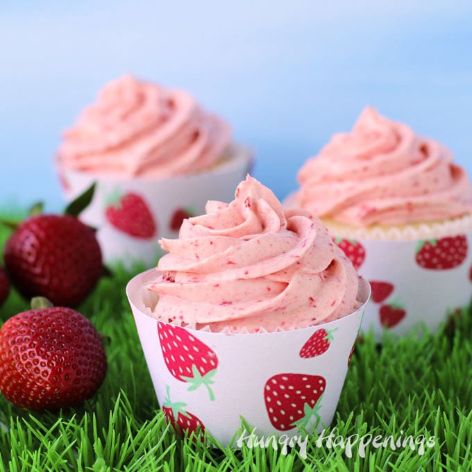 3 strawberry cupcakes wrapped in printable strawberry cupcake wrappers are set on grass in front of a blue sky background along with some fresh strawberries .