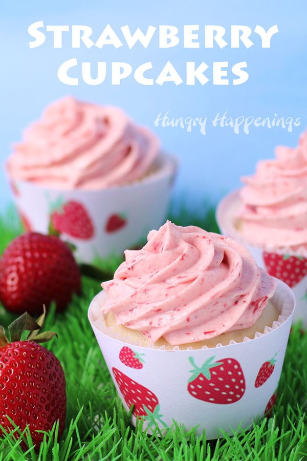 strawberry cupcakes wrapped in a strawberry printed cupcake wrapper are siting on plastic grass on a blue sky backdrop next to fresh strawberries