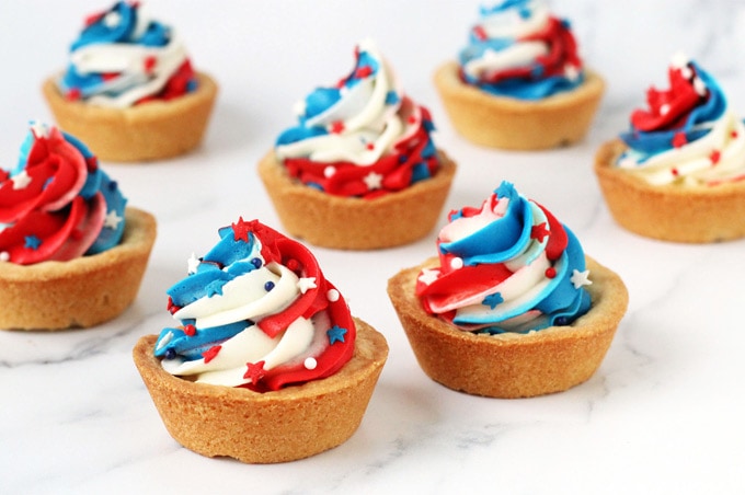 sugar cookie cups baked in a muffin tin are topped with a big swirl of red, white and blue frosting and star shaped sprinkles