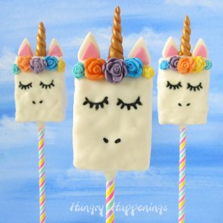 Three Unicorn Rice Krispie Treats on lollipop sticks are standing up in front of a blue sky backdrop