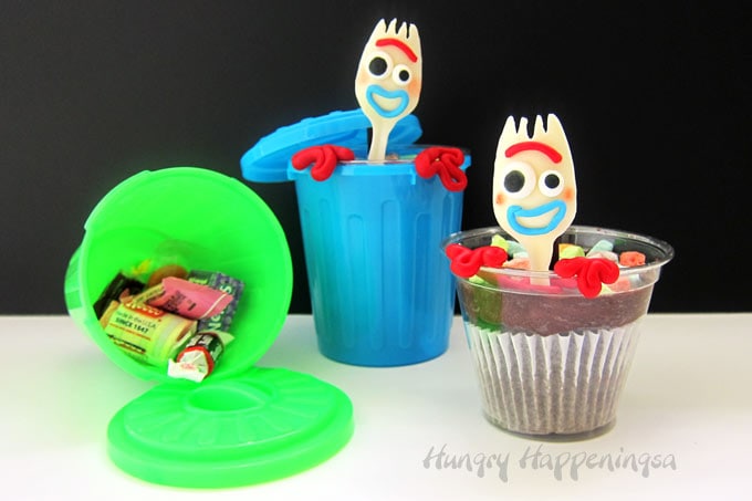 Green plastic trash can filled with candy is set next to a cupcake topped with a white chocolate Forky and both are set in front of a Forky Cupcake in a blue garbage can.