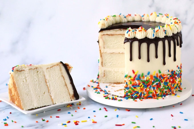 Sprinkle coated chocolate drip birthday cake cut open revealing the layers of white cake inside. 