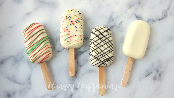 Four white cakesicles. One is plain while another is drizzled with red and green candy melts, another is topped with rainbow sprinkles, and another is drizzled with dark chocolate. 