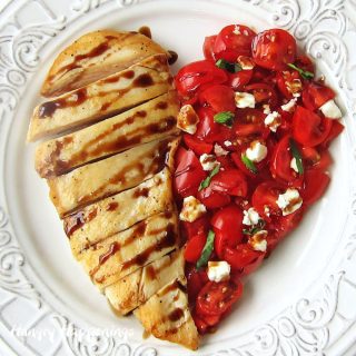 Chicken breast plated next to diced tomatoes, feta cheese, and basil to form a heart shape are drizzled with a balsamic glaze.