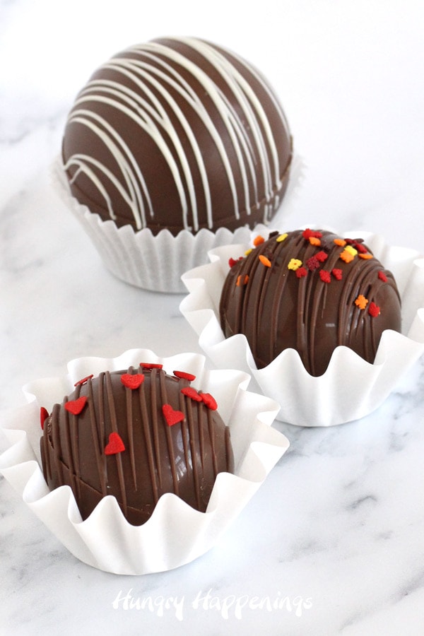 milk chocolate hot cocoa bombs decorated with red heart sprinkles, fall leaf sprinkles, and white chocolate drizzles