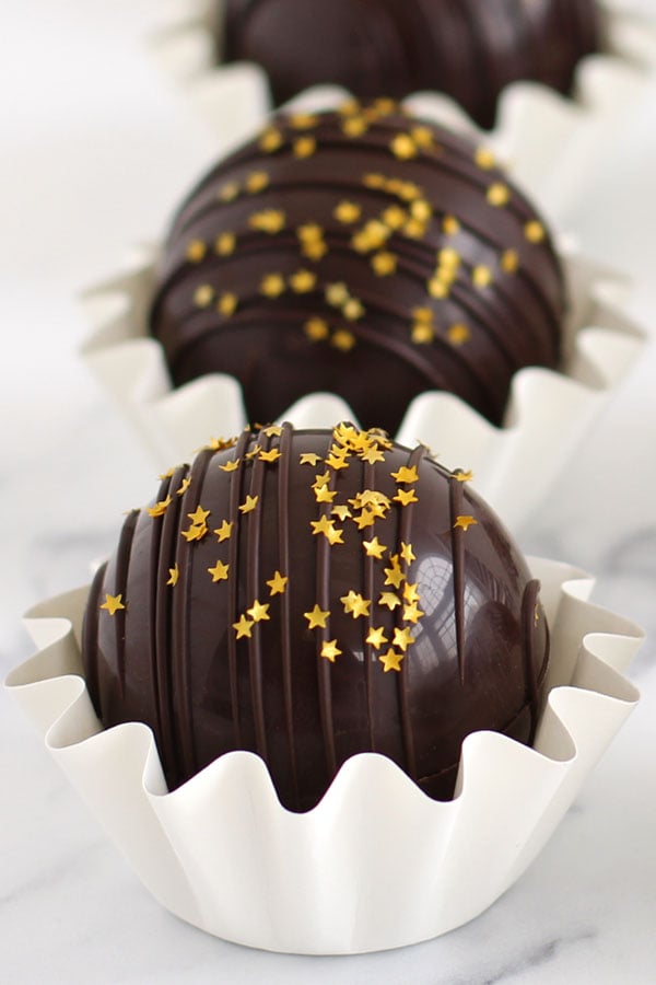 Hot chocolate bombs filled with liquor ganache are decorated with a drizzle of chocolate and edible gold star glitter