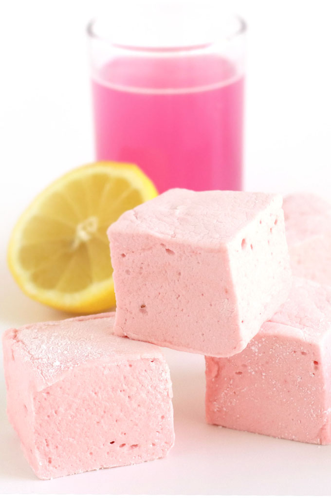 Strawberry Lemonade Marshmallows stacked in front of a glass of strawberry lemonade and a cut lemon.
