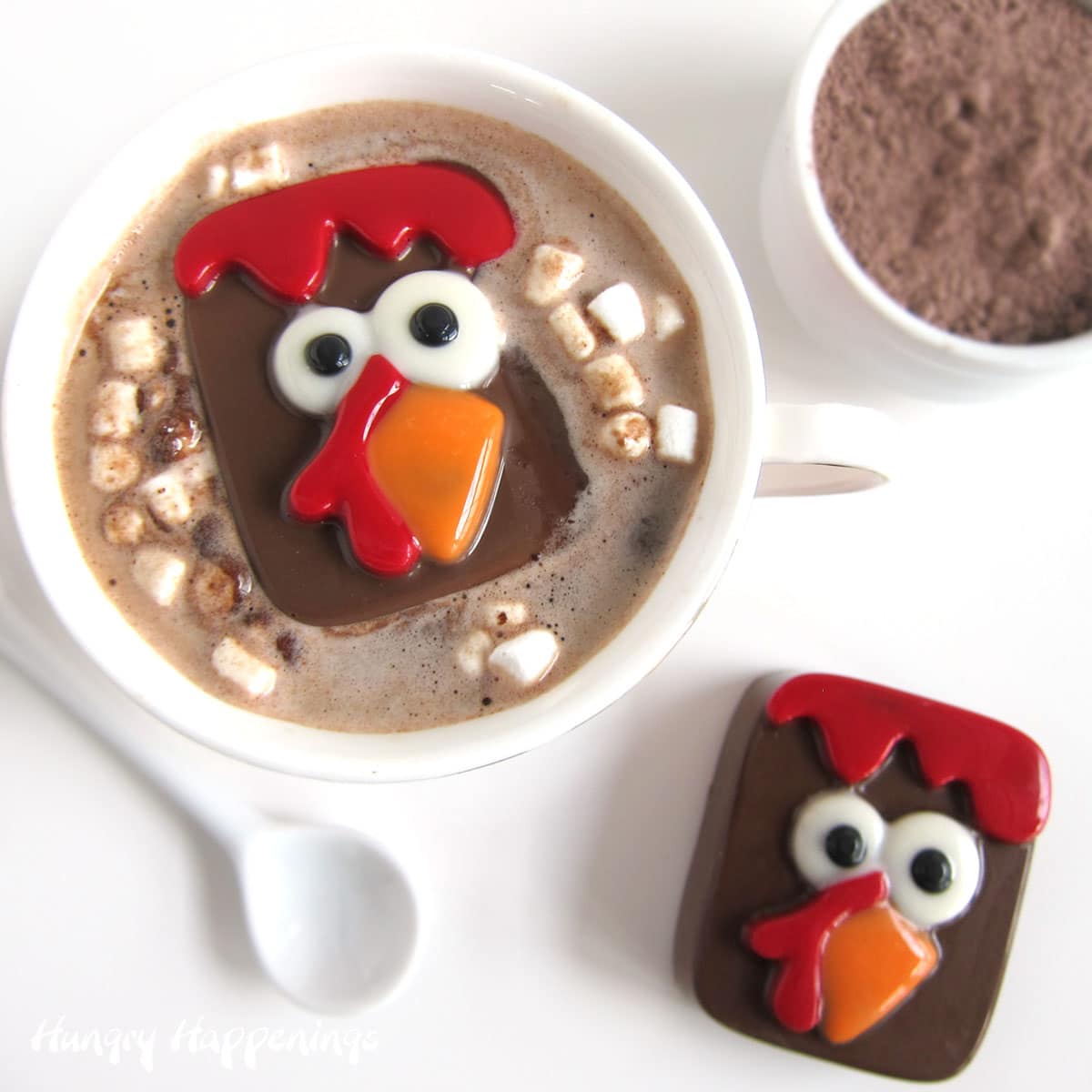 Turkey hot chocolate bomb in a mug of hot milk with marshmallows set next to a bowl of hot cocoa mix and a hot chocolate bomb turkey.