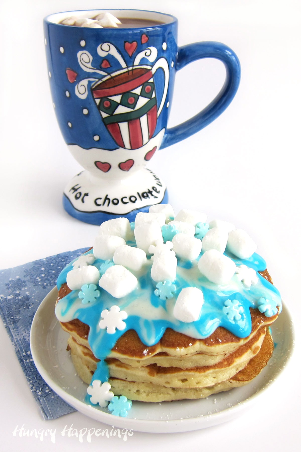 Winter Wonderland Pancakes topped with blue and white cream cheese icing, mini marshmallows, and candy snowflakes all served with a mug of hot chocolate.