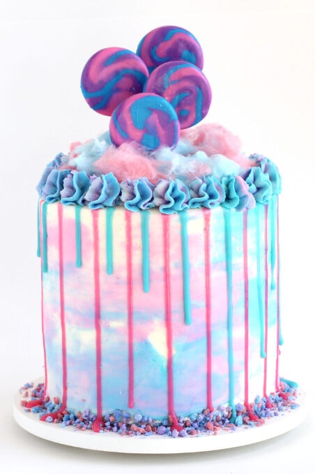 cotton candy drip cake frosted with pink, white, and blue icing and topped with cotton candy and lollipops