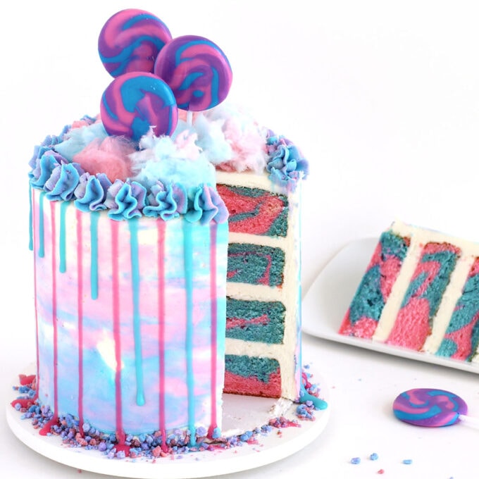 Pink and blue swirled cotton candy layer cake topped with drips of pink and blue ganache and fluffy cotton candy.