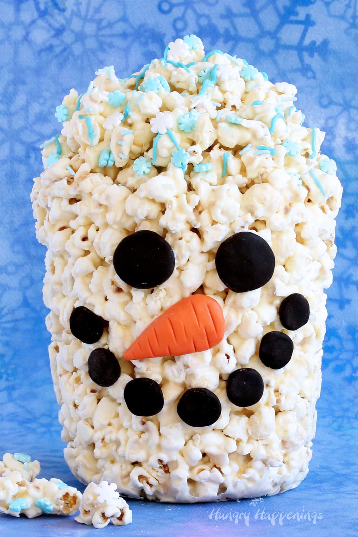 White chocolate popcorn bucket decorated like a snowman is filled with more popcorn sprinkled with snowflake candies.
