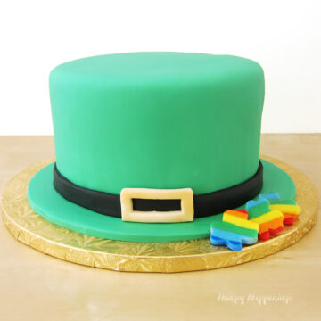 Leprechaun hat cake covered in green fondant set on a gold cake drum with rainbow shamrocks