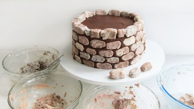 s'mores cake decorated with fudge stones set on a cake plate surrounded by bowls of cookies and cream fudge