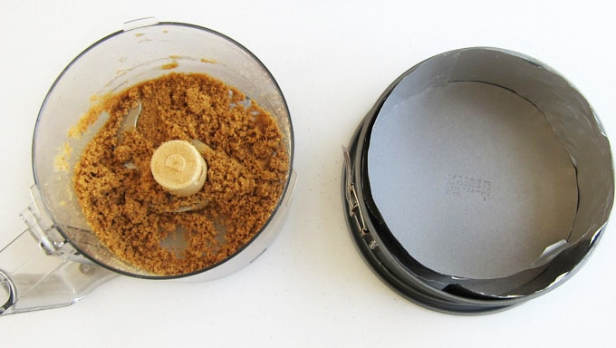 graham cracker crust mixture in a food processor bowl set next to a tin foil-lined springform pan
