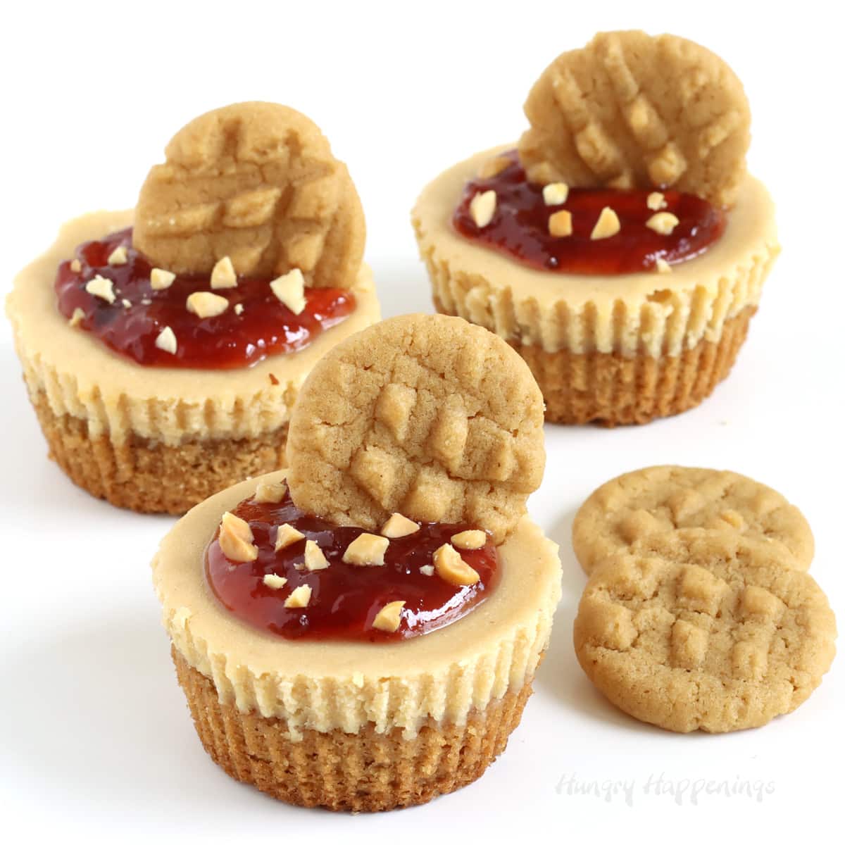 Mini peanut butter and jelly cheesecakes on peanut butter cookie crusts are topped with jam, chopped nuts, and mini cookies.
