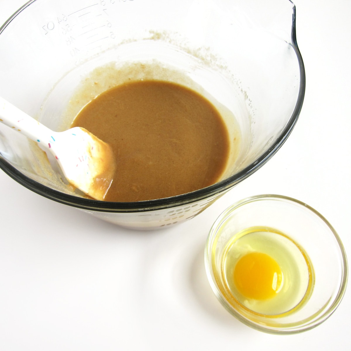 Mixing bowl with melted butter, peanut butter, brown sugar, and granulated sugar mixed together next to a ramekin with a cracked egg.