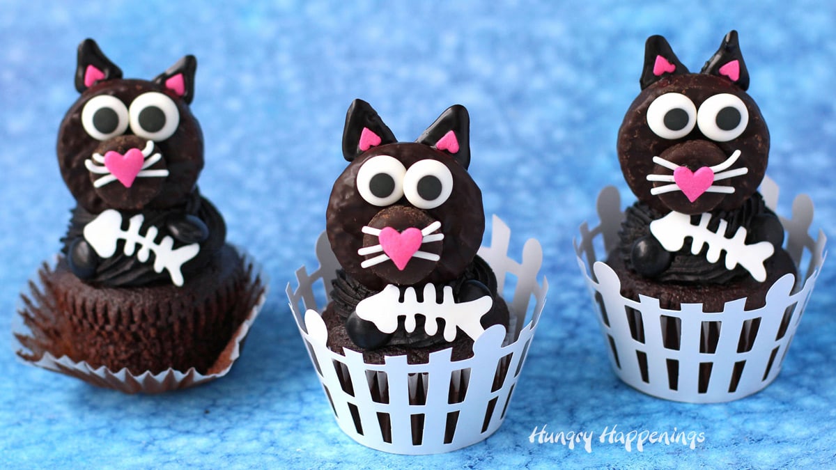 three black cat cupcakes in picket fence cupcake wrappers set on a blue watercolor background.