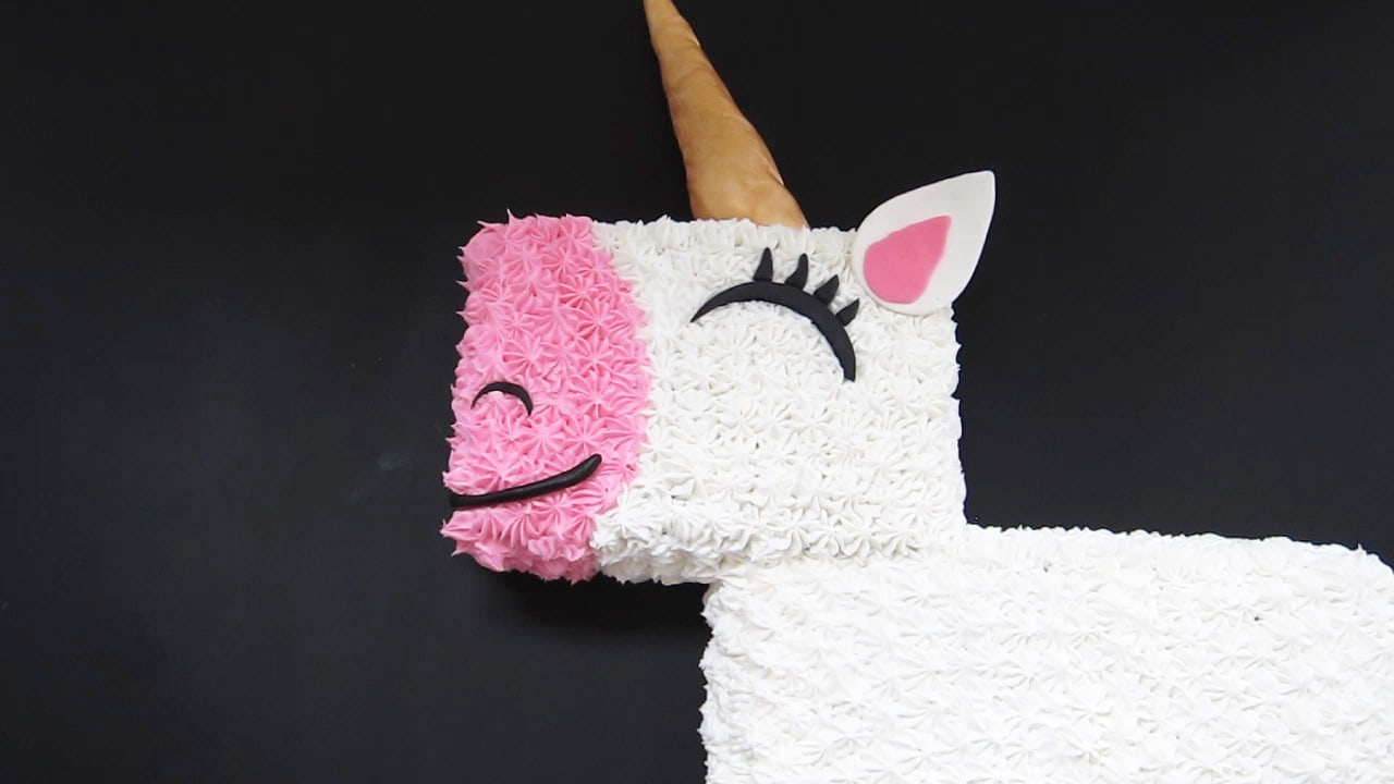 unicorn cake decorated with a gold modeling chocolate horn, a pink and white ear, and black eyes, mouth, and nose.