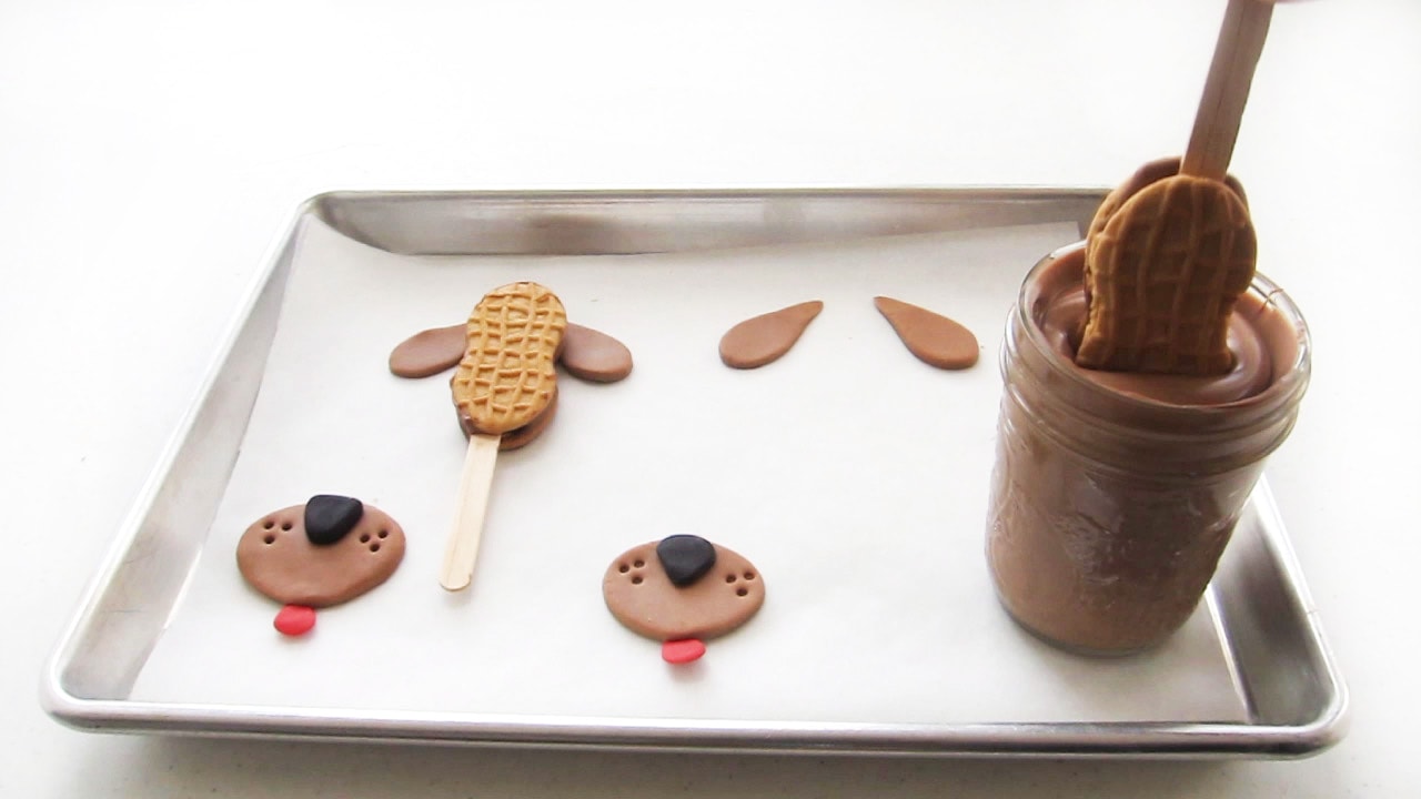 Dipping a Nutter Butter Cookie pop into a mason jar of chocolate that is set on a cookie sheet next to modeling chocolate dog features.