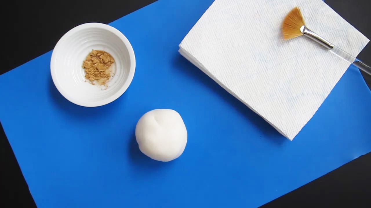 a ball of white modeling cholate on a blue silicone mat along with a bowl of gold luster dust and a fan paintbrush.