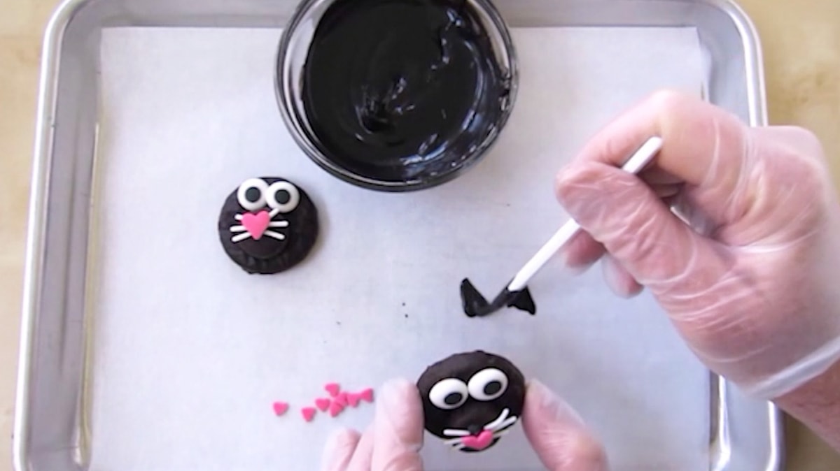 painting black candy melts ears onto a parchment paper lined baking sheet to attach the Peppermint Patty cat.