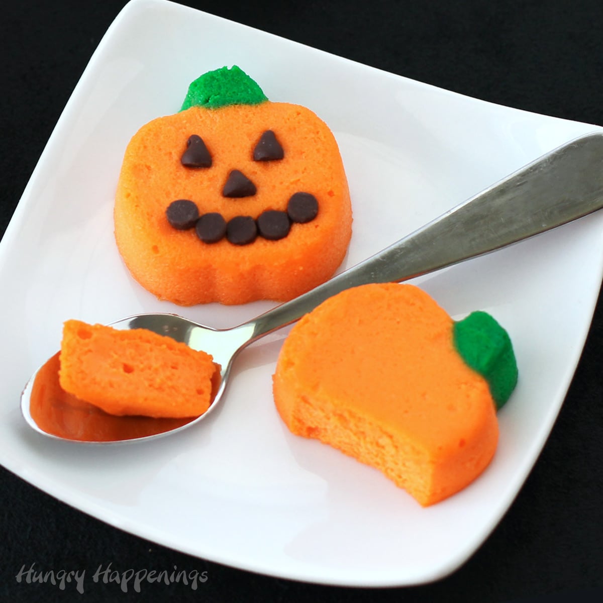 mini cheesecake pumpkins served plain or decorated with chocolate chips to look like a jack-o-lantern on a white square plate with a small spoon.