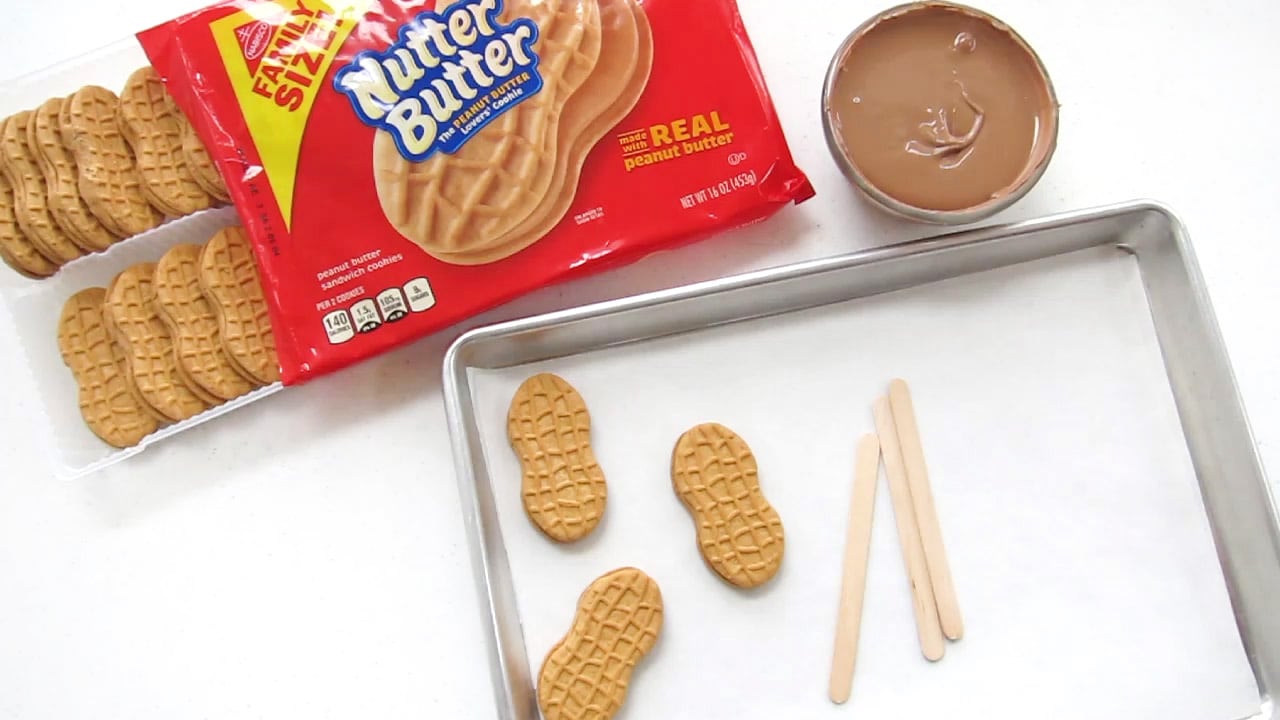 A family size package of Nutter Butter Cookies, a bowl of melted milk chocolate, and a tray of Nutter Butters and lollipops sticks.
