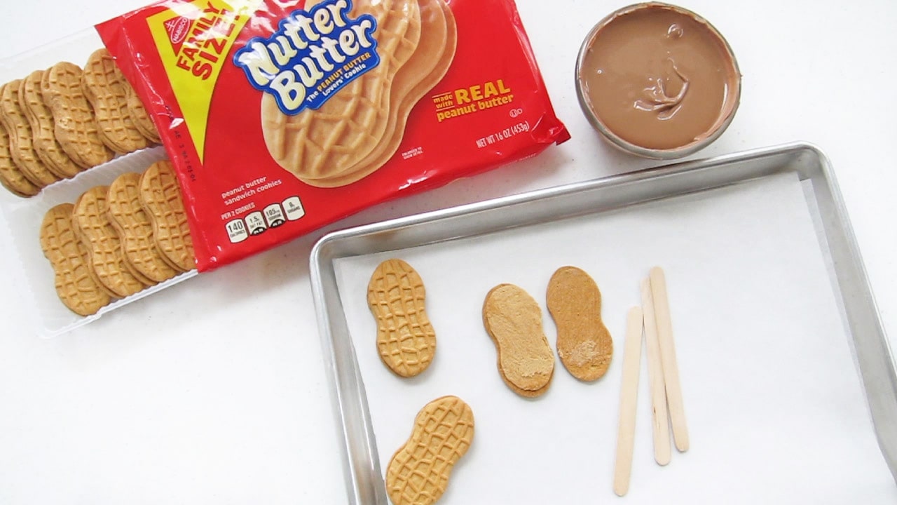 A Butter Cookie split open next to popsicle sticks on a baking sheet next to a package of Nutter Butters and a bowl of melted chocolate.