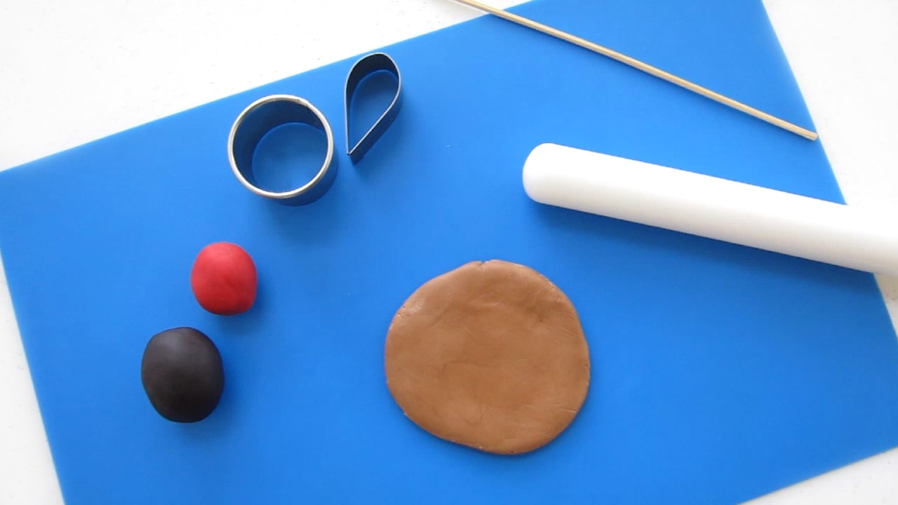 black, red, and brown modeling chocolate on a blue silicone mat with a round and teardrop cookie cutter, a fondant cutter, and wooden skewer.