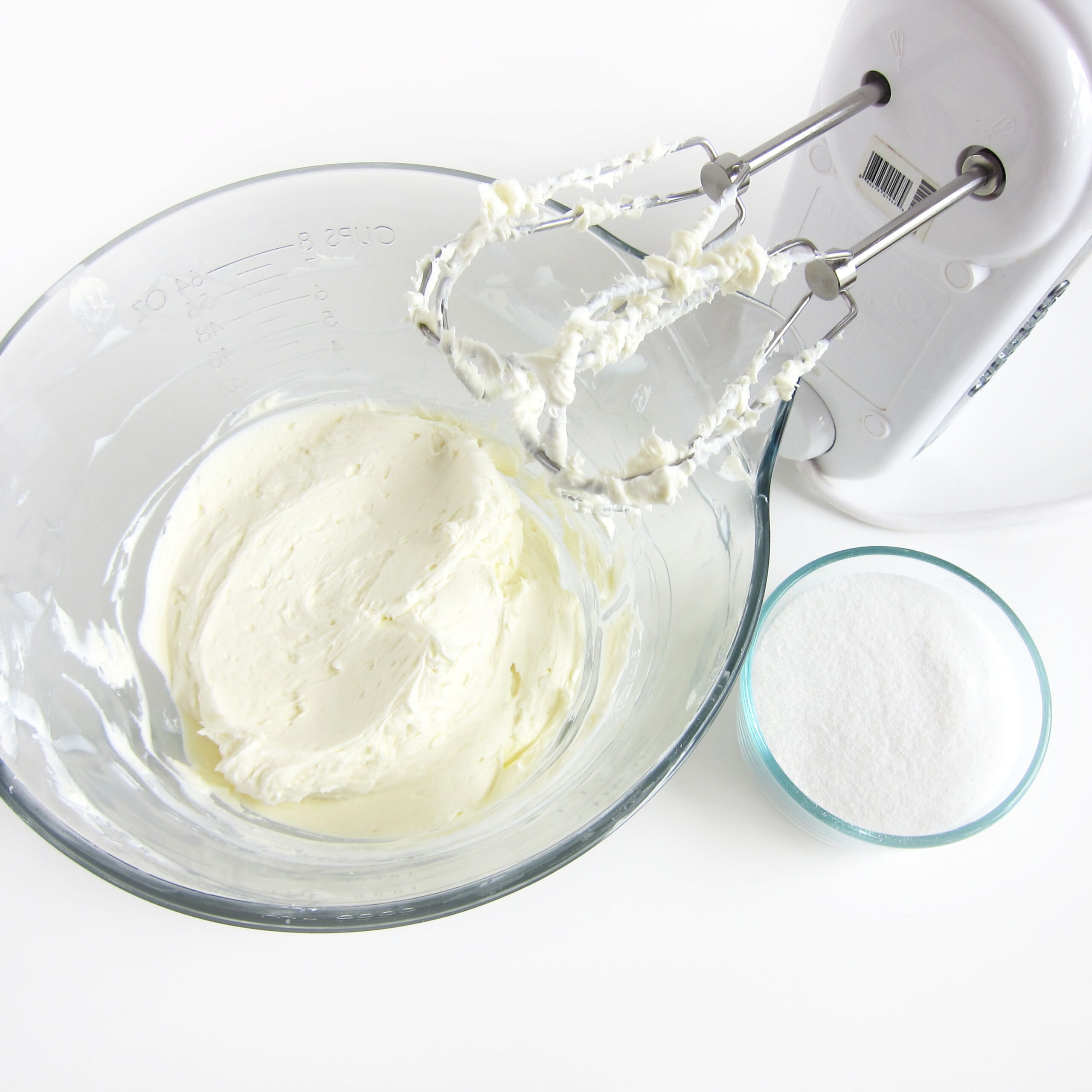 Cream cheese beaten until creamy in a mixing bowl next to a bowl of sugar and a mixer.