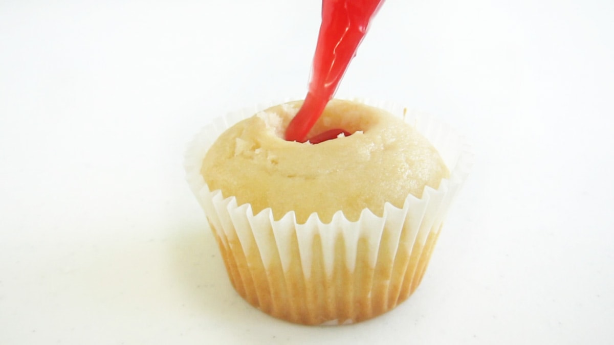 piping strawberry gel pie filling gel into the well in a white cupcake.