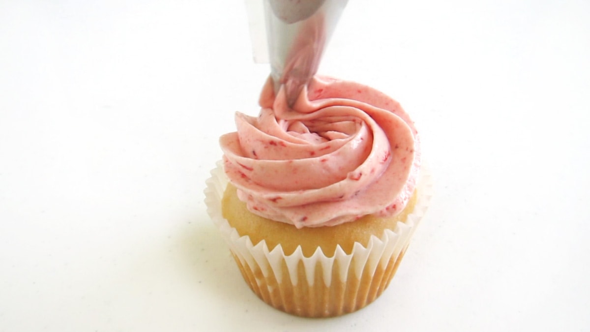 piping a swirl of strawberry frosting over a strawberry-filled white cupcake.