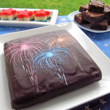 chocolate-glazed 4th of July brownie decorated with luster dust fireworks served on a white plate along with coconut cream candy stars and brownies.
