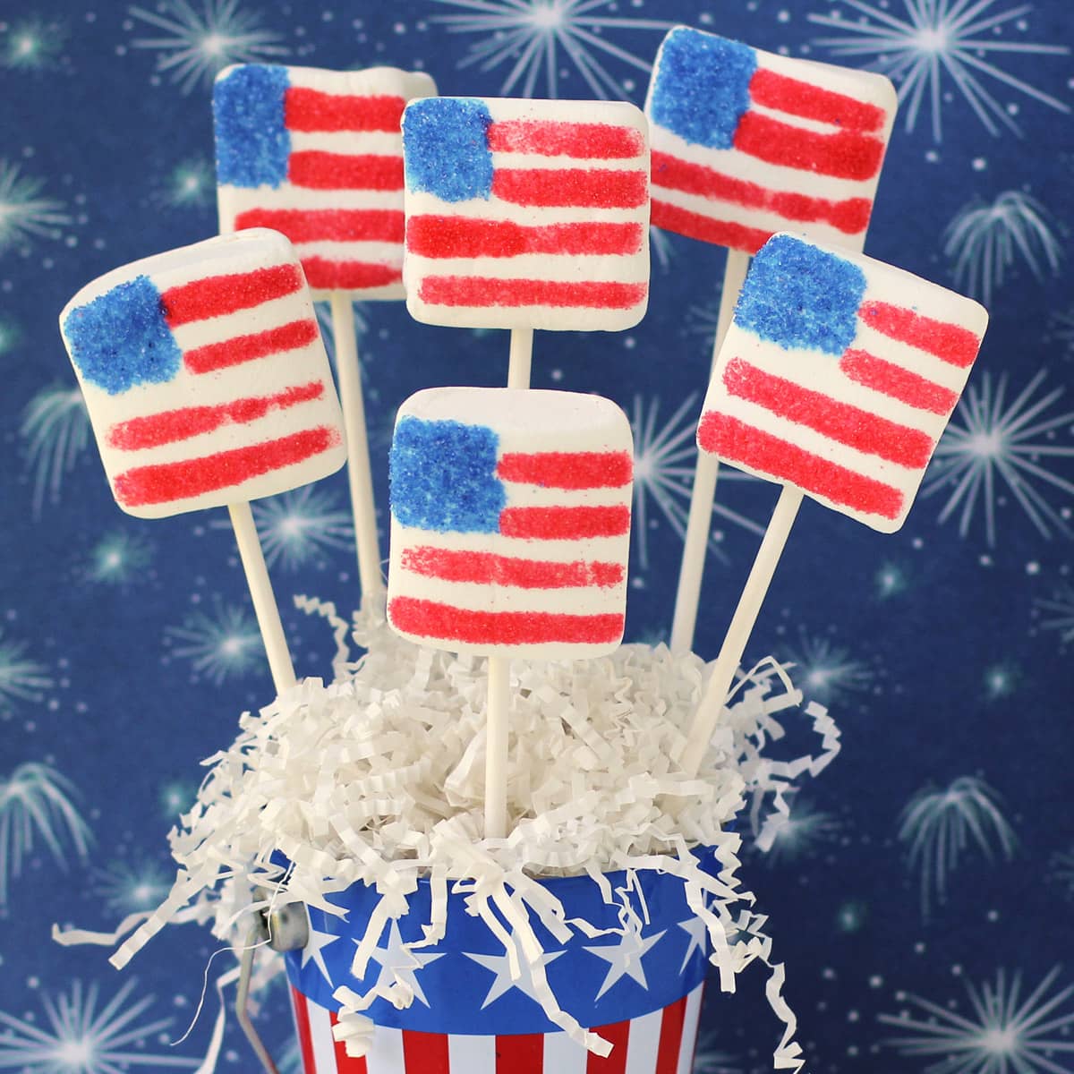 4th of July Marshmallow Flags on lollipop sticks arranged in a patriotic pail filled with white paper shreds displayed in front of fireworks paper.