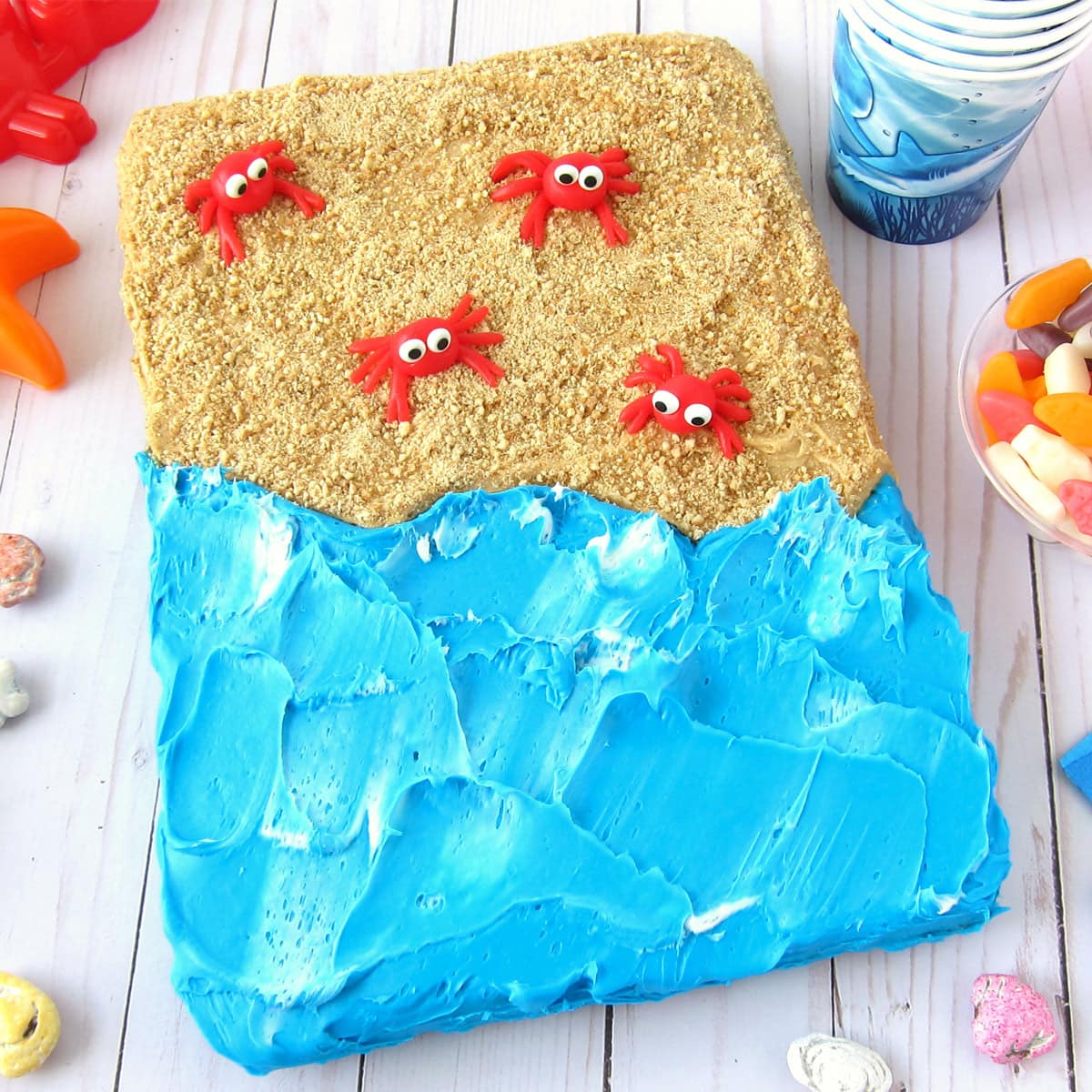 Beach cake made with a frosted brownie, blue frosting waves, cookie crumb sand, and candy crabs. Served on a table with candy shells, candy fish, and more.