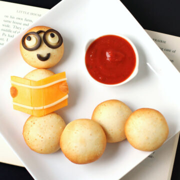 a Bookworm Snack decorated with cheese and olives in served with a bowl of marinara on a white plate set on a book.