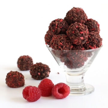 chocolate raspberry truffles coated in freeze-dried raspberries and chocolate shavings served in a glass candy dish next to fresh raspberries.