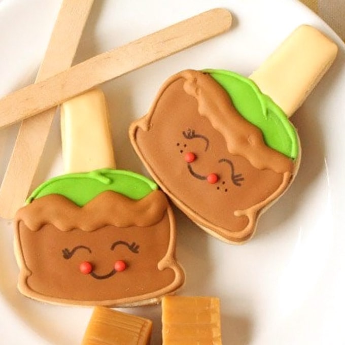 cute caramel apple sugar cookies with smiley faces on a white plate with popsicle sticks and caramels.