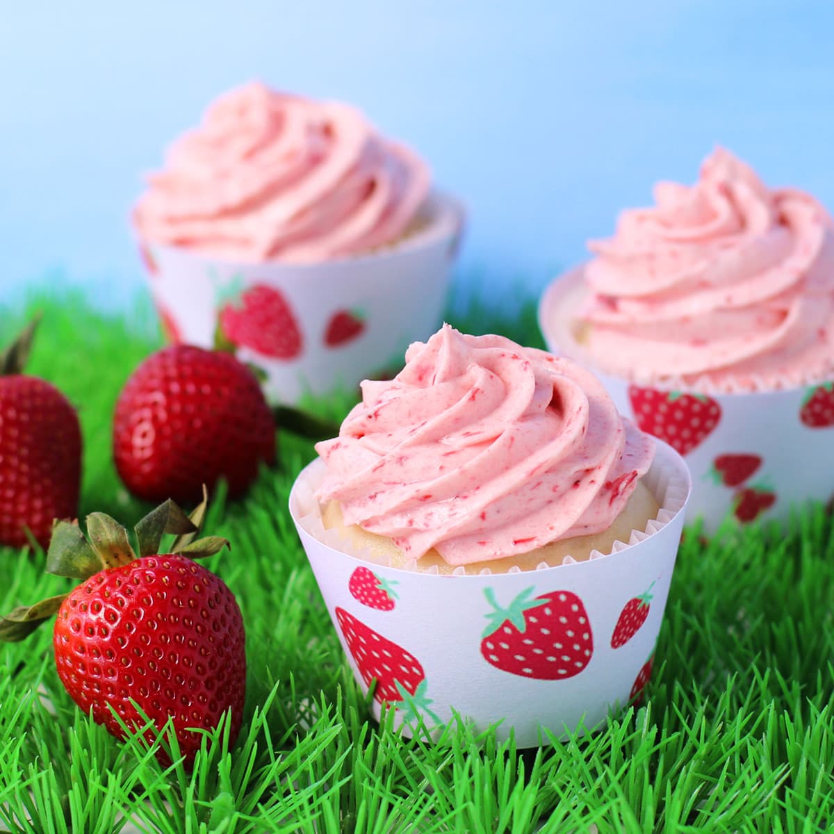 filled strawberry cupcakes topped with a swirl of strawberry frosting are wrapped in printed strawberry cupcake wrappers and are sitting in grass next to fresh strawberries.