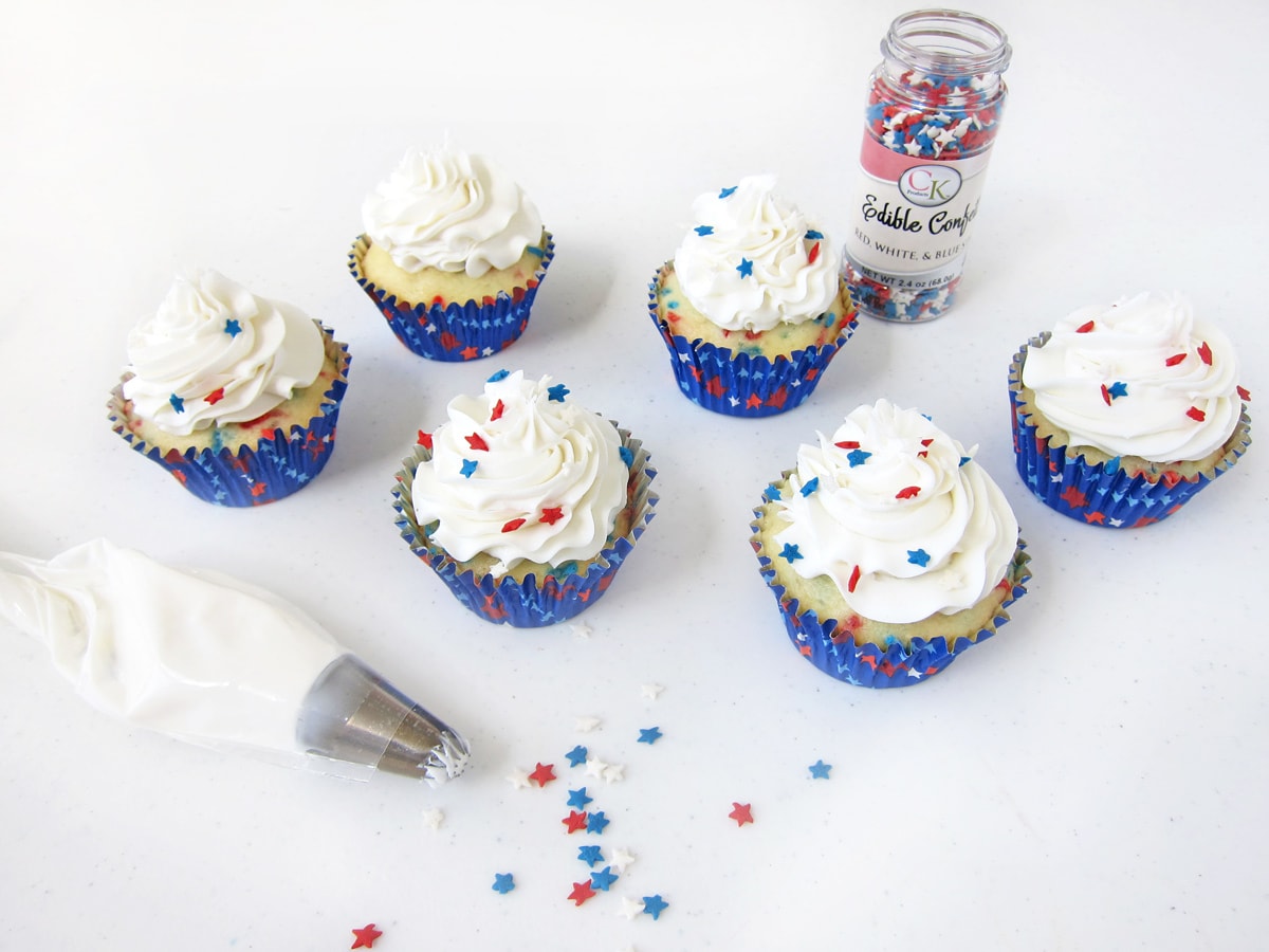 white funfetti cupcakes filled with red and blue sprinkles are frosted with a big swirl of white frosting and topped with red, white, and blue stars.