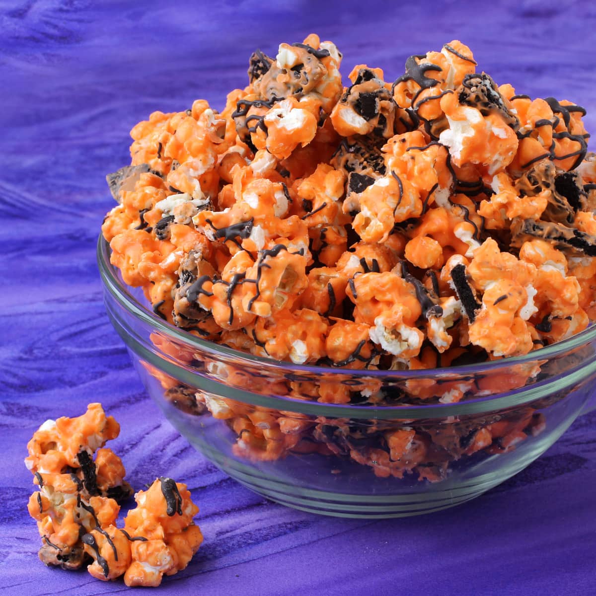 Orange and black cookies and cream Halloween popcorn served in a clear bowl on a purple watercolor table.