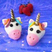 unicorn strawberries decorated with modeling chocolate sitting on a blue watercolor paper in front of fresh strawberries.