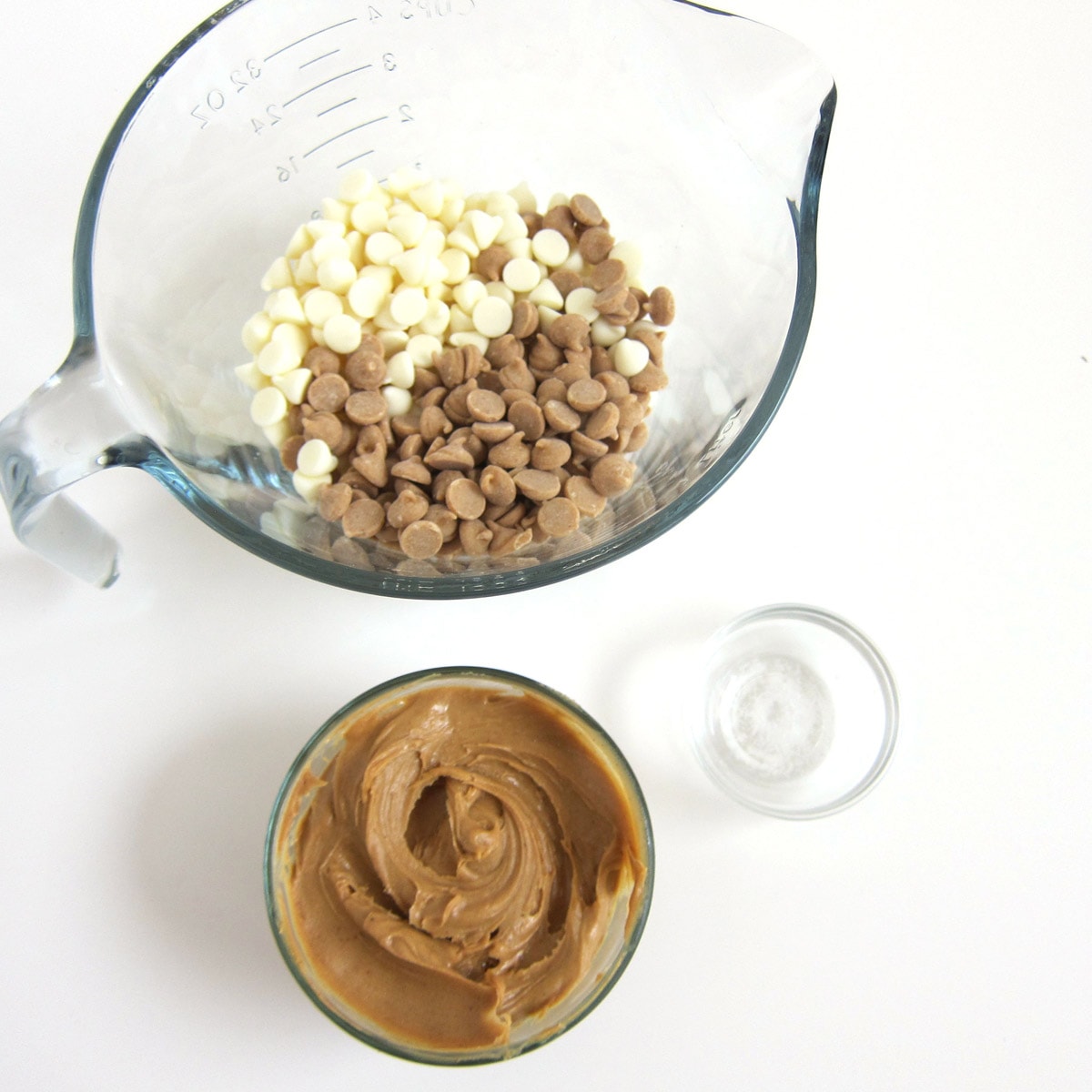 mixing bowl with white and peanut butter chips next to a bowl of creamy peanut butter and a bowl of salt.