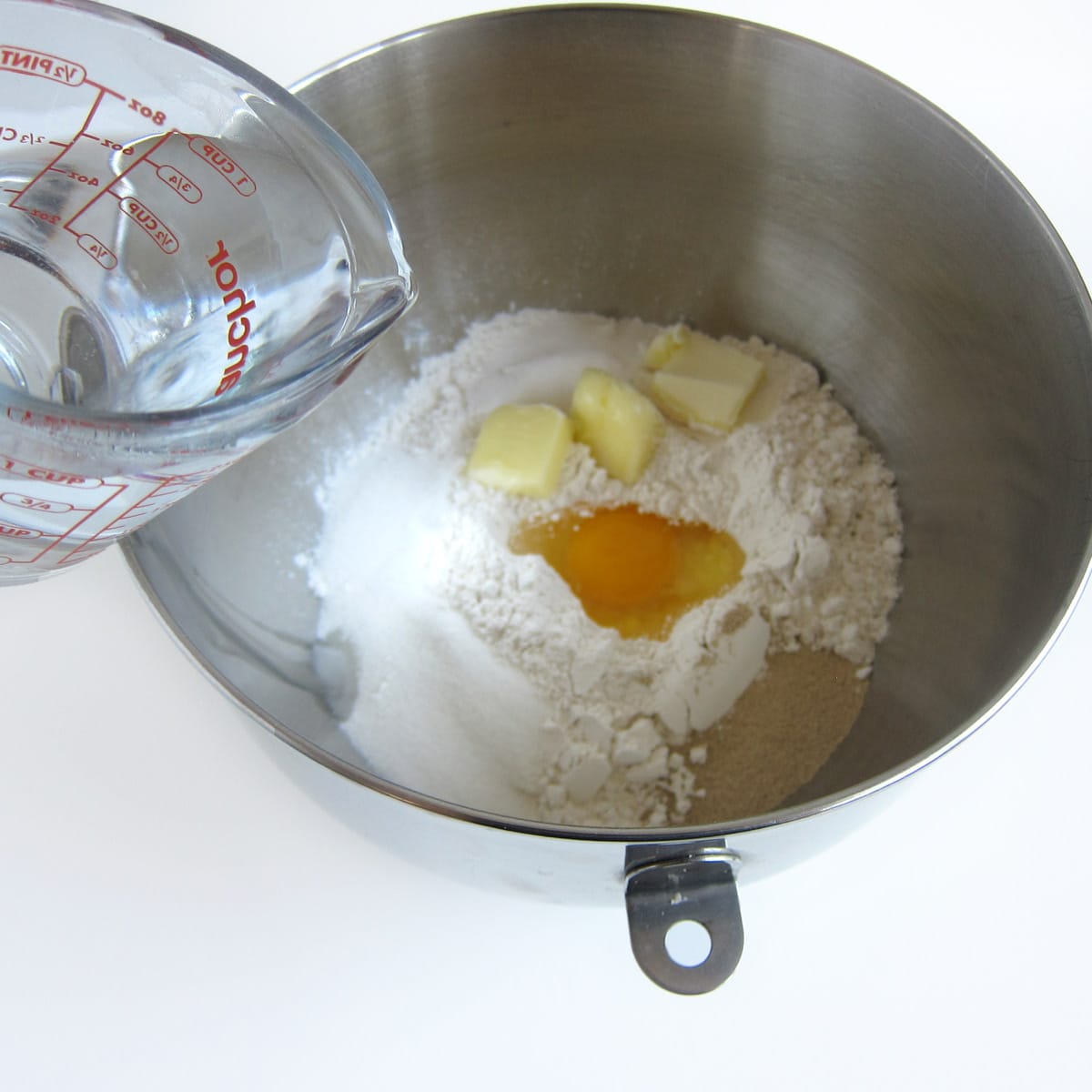pouring water into a bowl filled with flour, yeast, sugar, salt, egg, and butter.